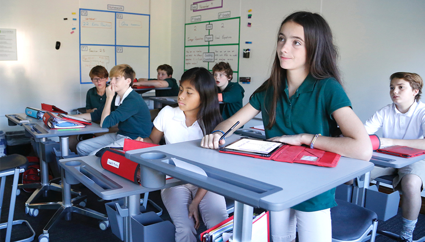 Young student listening to teacher at desk with her tablet.