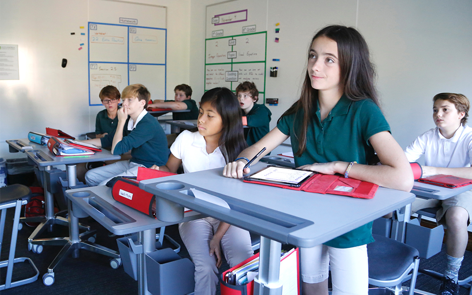 Young student listening to teacher at desk with her tablet.